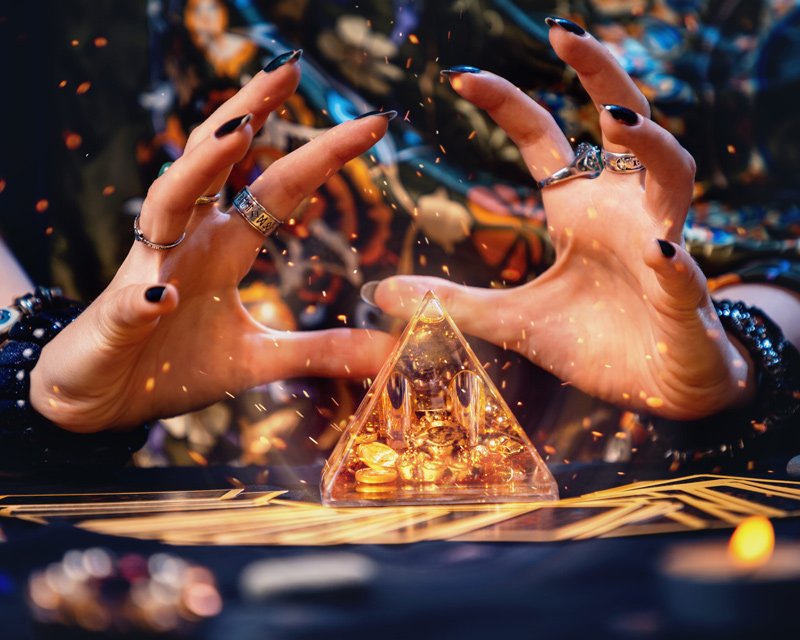 Close up of a psychic's hands around a sparkling gold pyramid. Gold sparkles swirl around the pyramid.